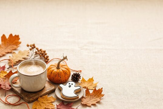 A cup of coffee sits next to a ghost-shaped cookie on a plate. A small pumpkin lies nearby on a table covered with autumn leaves. The setting captures a moment in fall