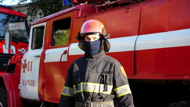 Portrait of male firefighter in helmet and balaclava standing near a big red car. Young confident fireman in full equipment looking into camera against the background of a fire truck. Close up