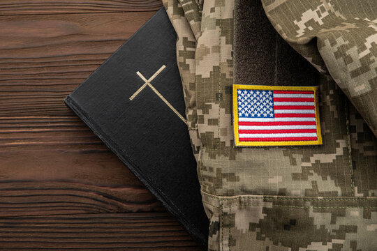 Christian cross on black book beside military camouflage uniform with American flag US, concept of faith and military service