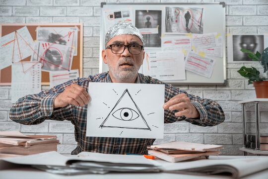 Worried conspiracy theorist in a tinfoil hat holding a hand drawn all seeing eye symbol in front of an evidence board.