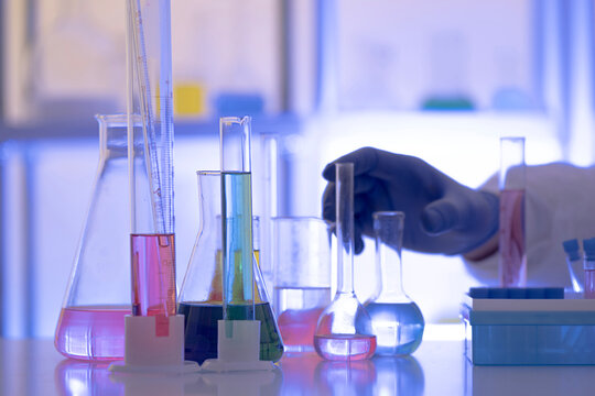 Chemistry laboratory scene with colorful chemical solutions in glass flasks and cylinders, scientist in gloves writing results, analytical experiment workspace.