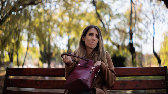 Stressed Young Woman Searching Her Handbag For Lost Item On Park Bench. Anxious female sitting on wooden bench outdoors in autumn trying to find missing objects inside purse