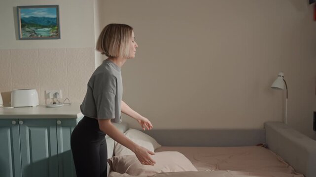 White woman fluffing pillow in bedroom, soft natural light, pastel bedding, calm smile, rhythmic movements suggesting music lover mood, casual grey top, bedside lamp, framed landscape painting