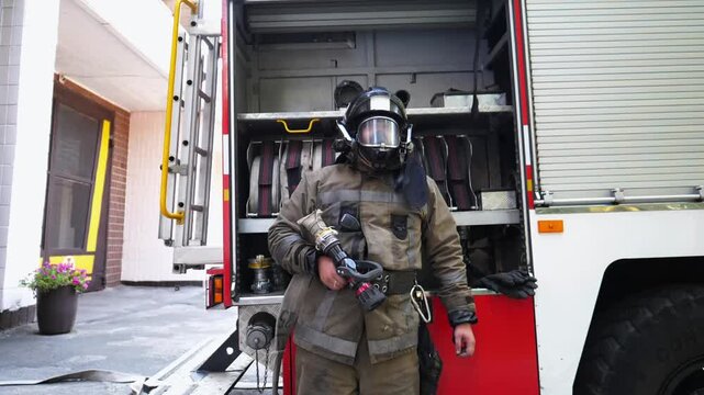 Portrait of young fireman in full equipment standing near a fire engine. Male firefighter with fire hose in hand looking into camera against the background of a big red car. Concept of saving lives