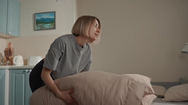 White woman stacking pillows and spreading sheet on bed, energetic rhythm in movement, cozy neutral decor, evening ritual feel, tidy linen, subtle music lover vibe, casual outfit, focused expression