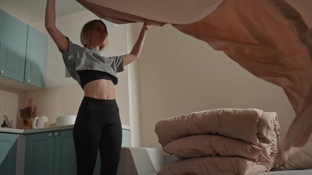 Young caucasian music lover making bed dancing to soft acoustic vibes in cozy kitchenette bedroom, smoothing pink sheets, casual crop top and leggings, warm sunlight, intimate calm mood, contentment