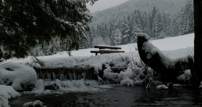 Mountain stream flowing over a small cascade next to snow-covered picnic benches and table in a winter forest