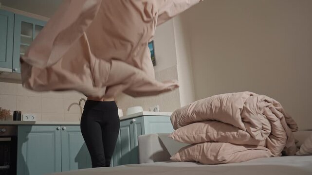 White woman dancing while making bed, music lover moving to upbeat playlist in bright studio apartment kitchen, tossing beige duvet with energetic motion, smiling in crop top and leggings, folded