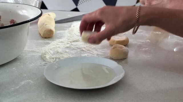 methodical hands rolling and shaping dough on kitchen countertop, surrounded by flour and plates, top-down view highlights concentrated preparation process, white flour and brown dough adda.