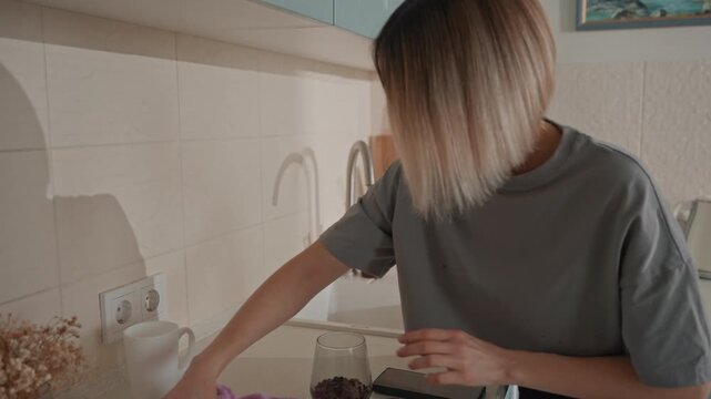 White woman wiping kitchen sink music lover cleaning with purple cloth ceramic mug on countertop utensils by backsplash calm reflective routine warm neutral tones shadow silhouette