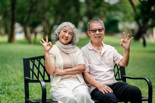 Joyful senior couple smiles at camera showing hand gestures on park bench for happy outdoor retirement
