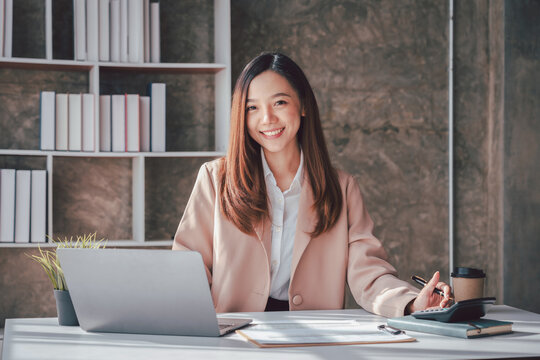 Smiling Asian Woman Working from Home Office with Laptop Calculator and Coffee Bookcase Background Achieving Financial Goals and Freelance Success