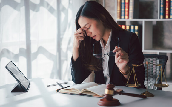 Stressed Asian Female Lawyer with Headache Feeling Exhausted at Work Legal Professional Burnout Pressure in Law Firm Office with Gavel and Scales of Justice