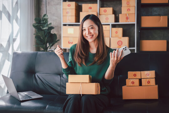 Asian Woman Managing Online Business with Smartphone and Laptop. Smiling Small Business Owner Checking Orders and Preparing Packages for Shipment Delivery at Home