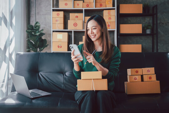 Young Asian Woman Managing Online Business on Sofa with Laptop and Boxes Home Office Setup Ecommerce Entrepreneurship and Parcel Delivery Service