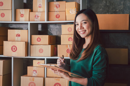 Asian Woman Managing Online Store and Checking Stock on Clipboard with Boxes. E-commerce Business and Dropshipping Logistics for Online Retail Delivery Service