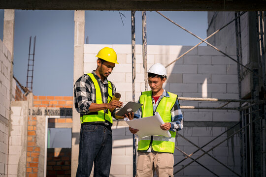 Two Asian Construction Workers Reviewing Blueprints and Using Tablet at Construction Site Checking Progress on Building Project