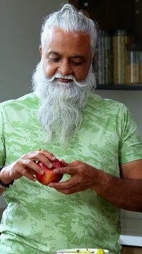 Indian mid age man holding and showing fresh red apple to camera, stylish bearded male promoting healthy eating while flexing bicep muscle and pointing at fruit standing in modern kitchen interior