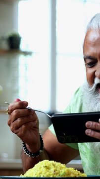 Indian mid age man watching smartphone reels while eating at home, stylish bearded male enjoying poha at breakfast table and streaming web series or comedy videos in modern kitchen lifestyle