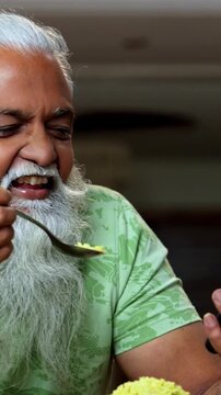 Indian mid age man watching smartphone reels while eating at home, stylish bearded male enjoying poha at breakfast table and streaming web series or comedy videos in modern kitchen lifestyle