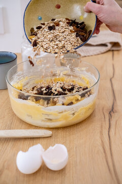 Homemade breakfast process. Healthy recipe. Female chef hand pouring oat flakes raisins in dough bowl on kitchen counter with eggshell.