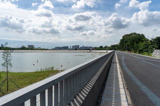 Long road beside expansive city lake under cloudy sky