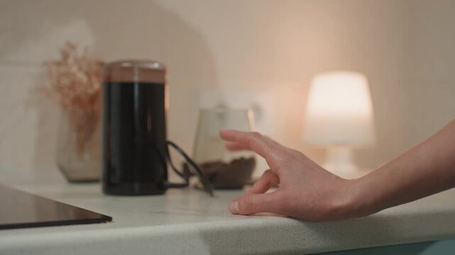 Hand on kitchen counter near speaker, warm lamp glow, corded device and glass in background, fingers hover then press edge of countertop, latenight music lover reaching for audio device, intimate cozy