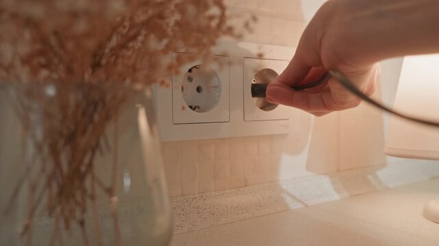 Cozy bedside hand plugging lamp. Warm ambient light casts soft shadows over ceramic vase with dried flowers, textured tabletop and double socket. Slow deliberate motion of hand inserting plug into