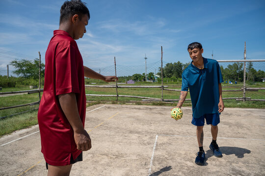 Asian man holding takraw ball preparing to serve to teammate on outdoor court. Two men standing on a concrete court preparing to start a casual game of sepak takraw on a sunny afternoon