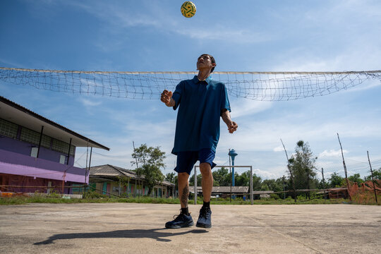 Asian man looking up to head sepak takraw ball over net on outdoor court. Athletic man in blue sportswear focusing on a high-floating takraw ball during a game on a sunny outdoor court.