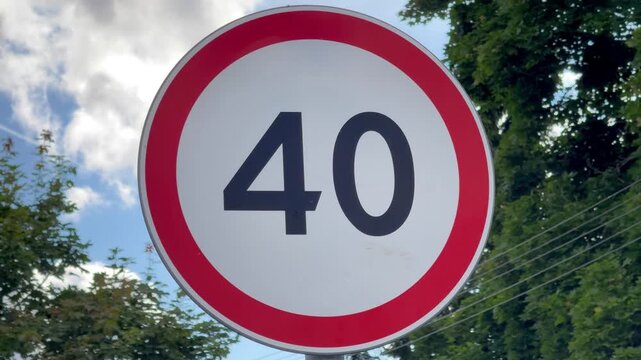 Round speed limit 40 road sign with red border against blue sky with clouds and green trees, traffic sign, speed limit, road safety, urban transport.