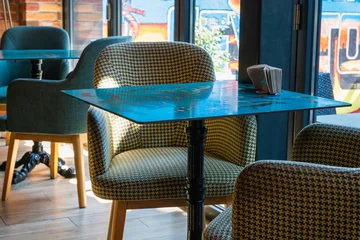 Fotobehang Horeca A cozy cafe interior featuring shiny blue square tables surrounded by stylish, patterned upholstered armchairs bathed in natural light near a window.  © Andrey