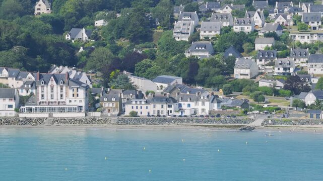 Aerial view of the coastal town of Binic in Brittany, France, showing traditional houses clustered on a green hillside above a sandy beach and turquoise sea.