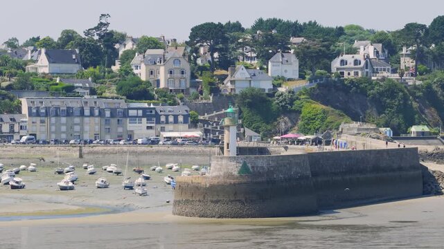 Wide shot of Binic port , showing a stone pier with a green-topped lighthouse, numerous small boats resting on the sand at low tide in Brittany, France.
