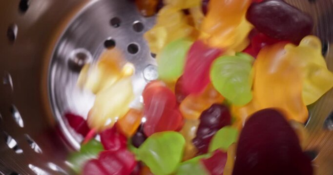 Slow motion tumbling of colorful gummy candies inside stainless steel colander into industrial conveyor, Food production.