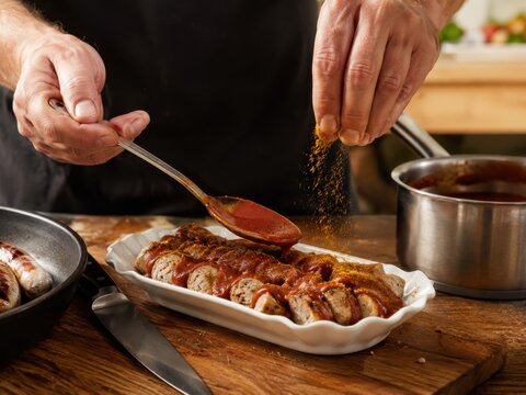 Currywurst wird frisch angerichtet: H&auml;nde geben w&uuml;rzige Tomatenso&szlig;e und Currypulver &uuml;ber gebratene Bratwurstst&uuml;cke &ndash; ber&uuml;hmter deutscher Imbissklassiker und beliebtes Streetfood.