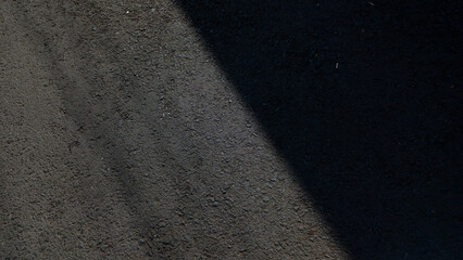 Fototapeta premium Dark rough asphalt road surface texture split by diagonal sunlight shadow.