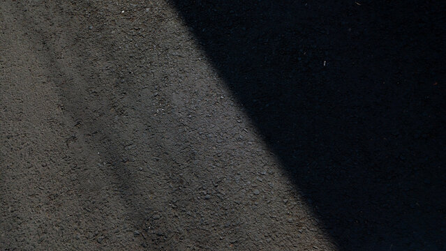 Dark rough asphalt road surface texture split by diagonal sunlight shadow.