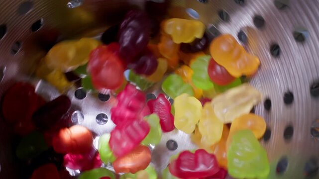 Slow motion gummy candies tumbling inside stainless steel colander into industrial conveyor, Food production.