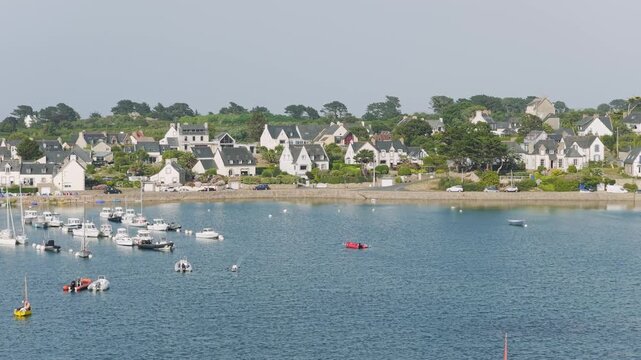Wide daytime view of the entrance to the port of Ploumanac'h in Perros-Guirec, Brittany, France, calm blue water with anchored boats and yachts, a shoreline lined with traditional Breton houses.
