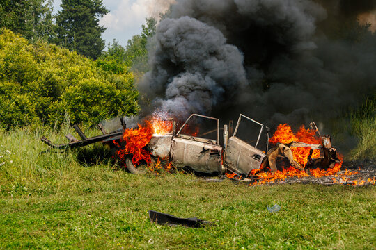 Old car burning in a green grassy field with bright orange flames and thick black smoke rising into the sky against a background of trees and bushes on a summer day