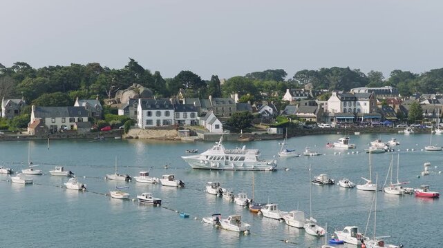 A wide view of the entrance to the port of Ploumanac'h in Perros-Guirec, Brittany, France, showing numerous small boats and a larger vessel anchored in calm blue water.