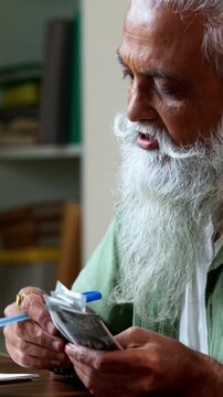 Indian mid age man counting Indian money notes at home, stylish bearded male managing monthly finance using calculator and diary while handling five hundred rupee currency notes at table