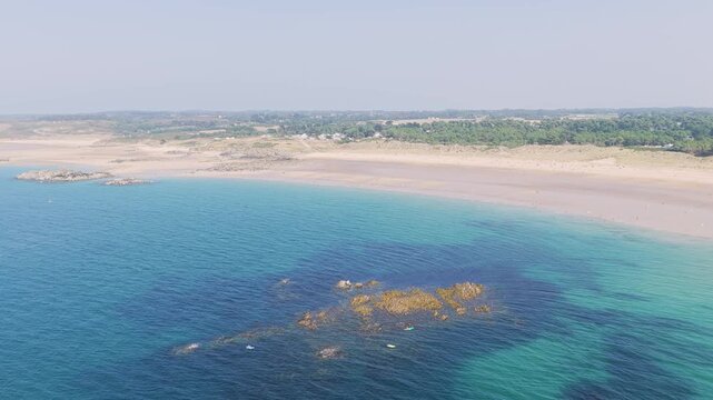 Aerial view of Anse du Croc beach in Frehel, Brittany, France, showing turquoise waters, rocky outcrops, and a wide sandy shoreline under clear summer skies.