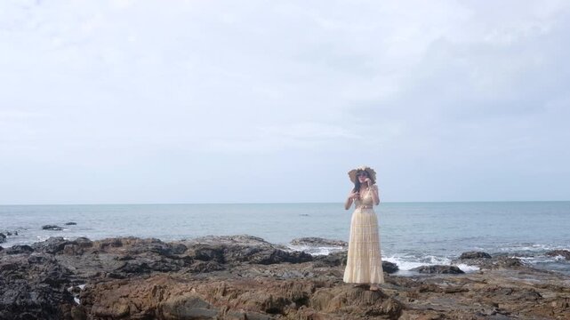 Young Asian Woman in Maxi Dress and Straw Hat Standing on Rocky Ocean Shore