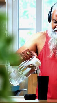 Indian mid age fit man with beard making protein shake post workout at home, stylish healthy male wearing headphones and sando vest preparing nutrition supplement in modern kitchen interior