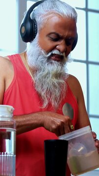 Indian mid age fit man with beard making protein shake post workout at home, stylish healthy male wearing headphones and sando vest preparing nutrition supplement in modern kitchen interior