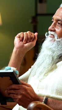 Indian mid age bearded man holds photo frame while sitting on rocking chair at home, stylish male with wet eyes recalling memories of loved one in modern interior emotional lifestyle moment