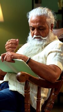 Indian mid age bearded man reading book novel on relaxing chair at home, stylish mature male enjoying peaceful leisure time with printed copy in modern interior lifestyle setting
