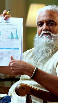 Indian mid age bearded man reading and showing financial document to camera sitting on relaxing chair at home, stylish mature male evaluating profit and loss report with positive expression